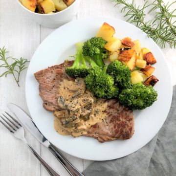 Rose veal escalope with creamy mushroom sauce and rosemary roasted potatoes and broccoli on plate with roasted potatoes in bowl to side with fresh rosemary leaves
