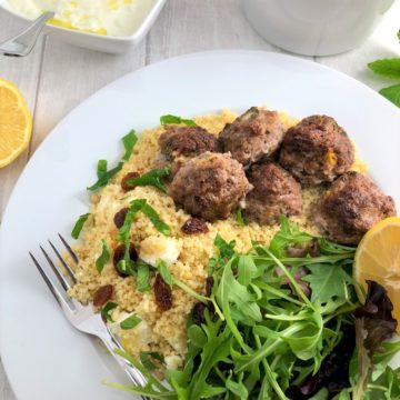 lamb meatballs served on bed couscous with apricots and sultanas and green salad and lemon with bowl greek tzatziki to side and bowl couscous to side