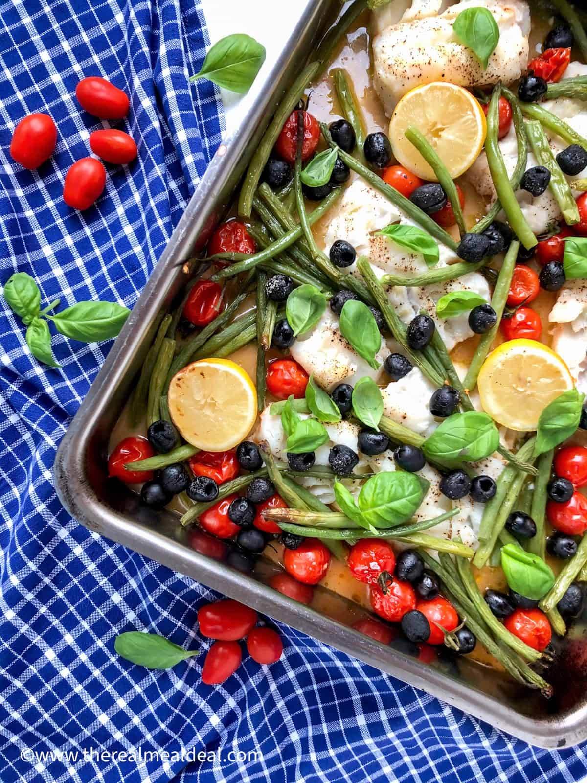 baked cod black olives cherry tomatoes and green beans in metal baking tray topped with fresh basil leaves