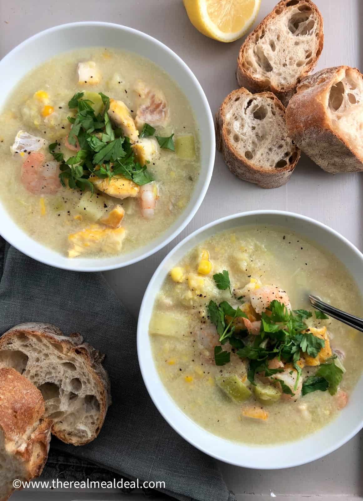 two bowls of smoked haddock and prawn chowder on a tray with sliced baguette half lemon and bowl of chopped parsley