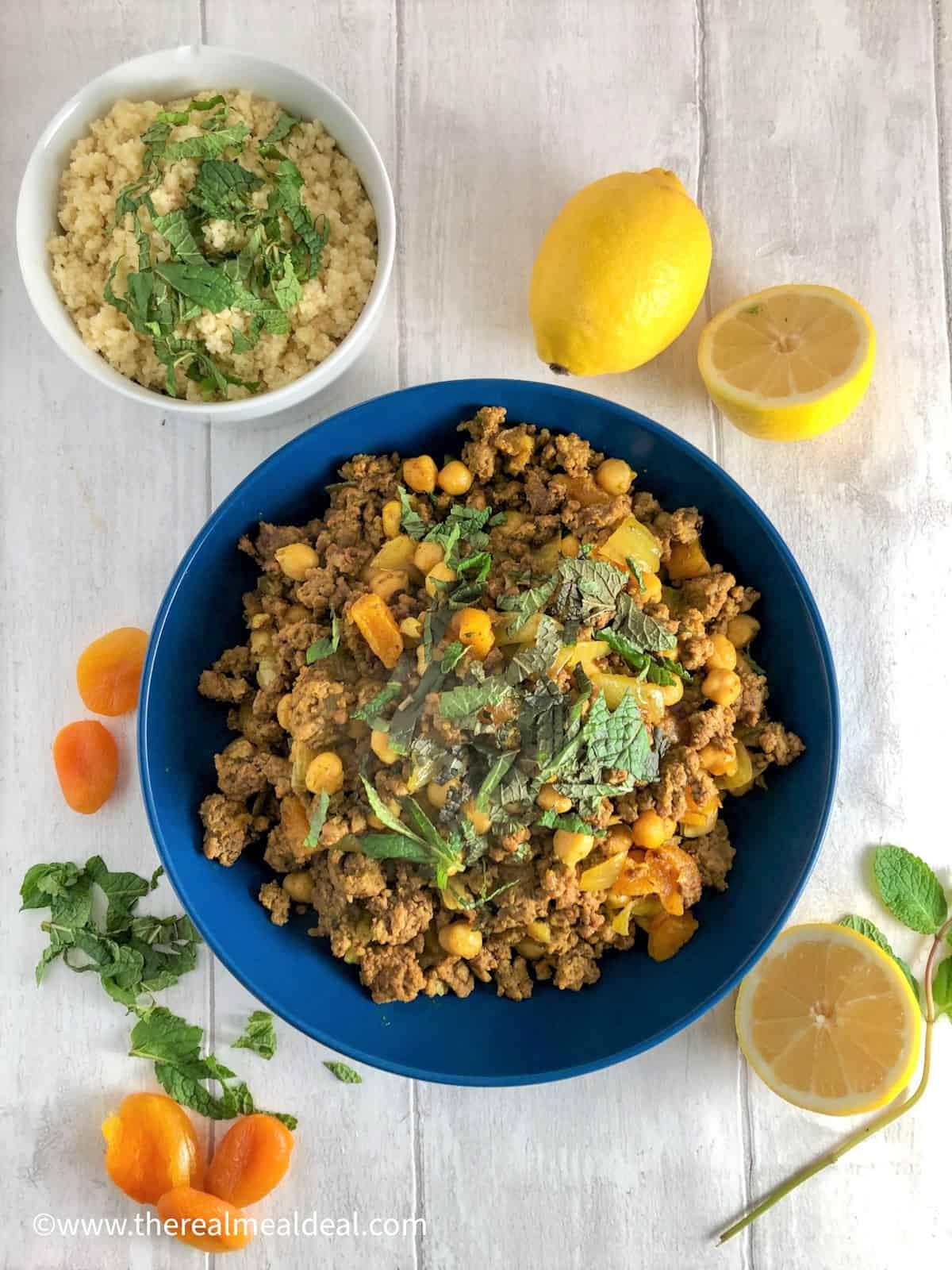 Lamb tagine with apricots and chickpeas in a bowl topped with fresh mint and dried apricots and lemon halves to the side and a bowl of couscous