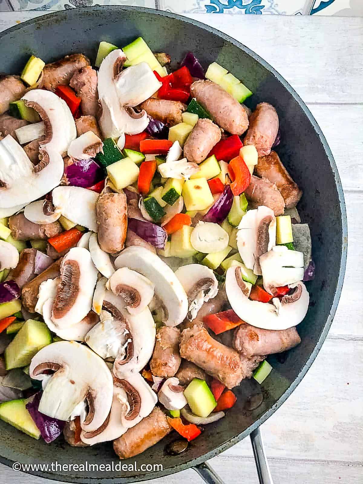 zucchini/courgette, mushrooms, peppers, red onion, sausages frying in pan.