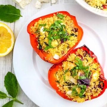two red pepper halves stuffed with vegetable couscous and feta cheese on plate with lemon, mint and feta cheese to side