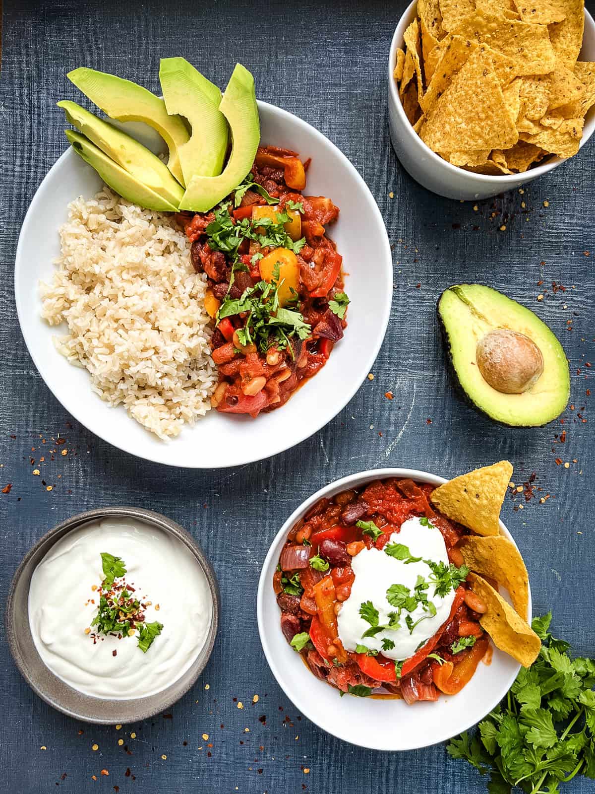 bean chilli in bowl topped with soured cream with sides of avocado soured cream and bowl of chilli with rice