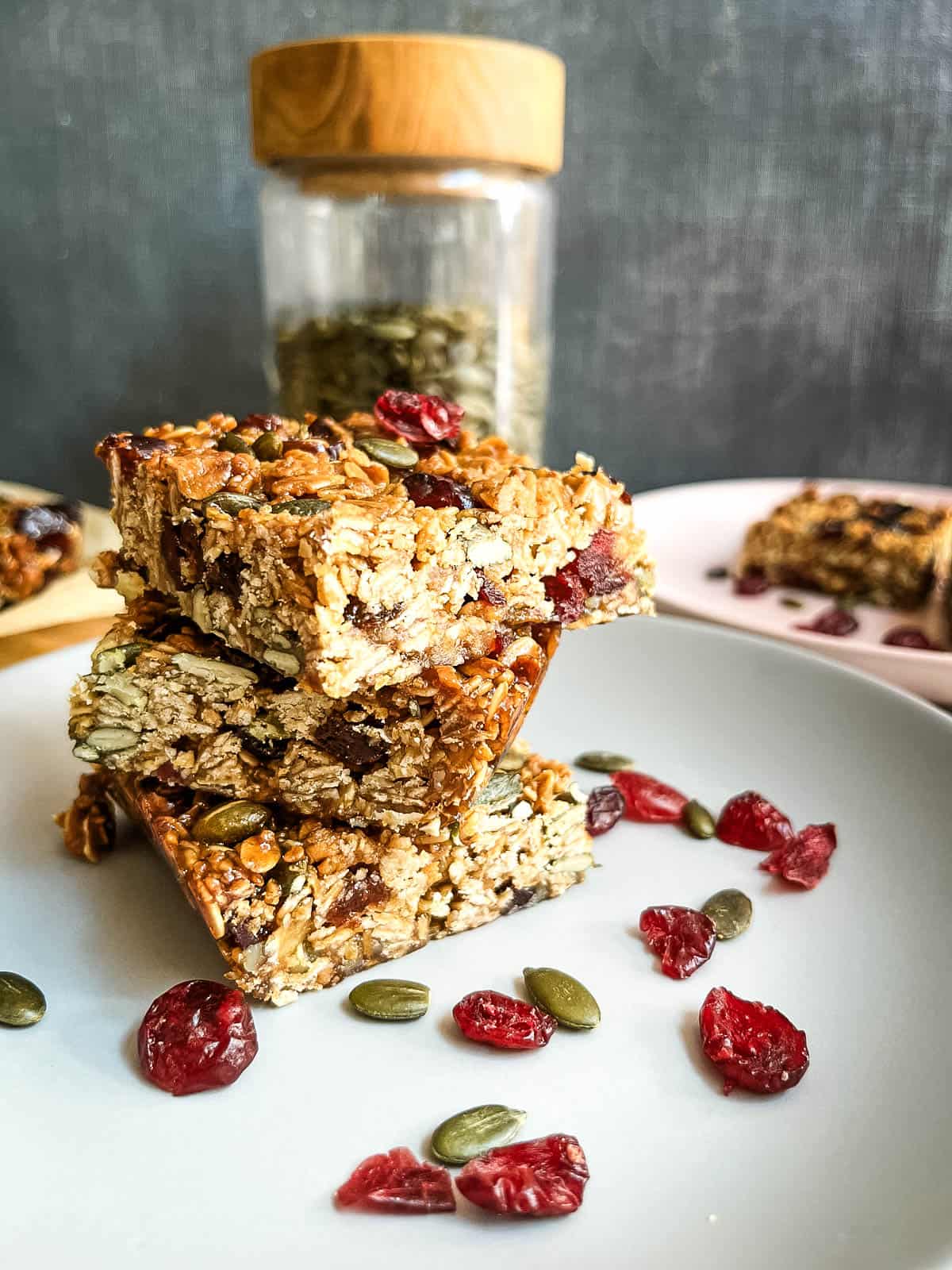 3 granola bars stacked on plate with dried cranberries and pumpkin seeds.