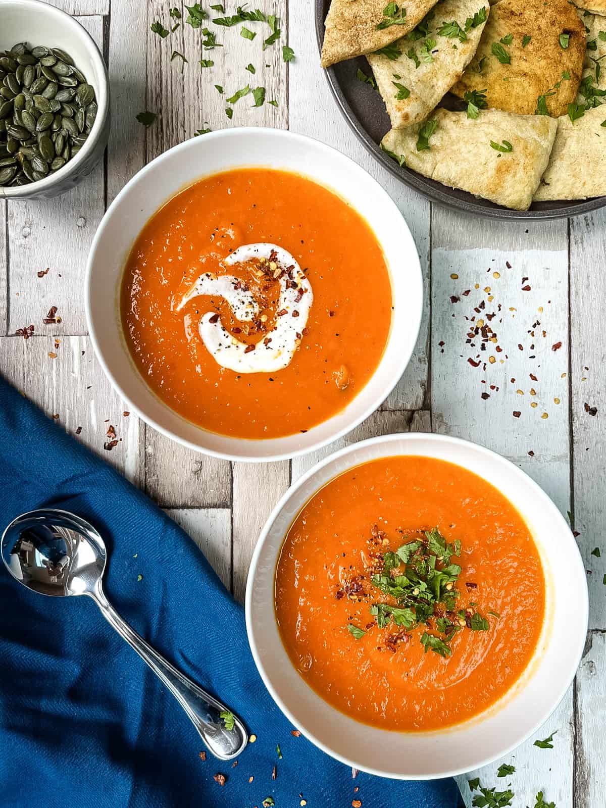 two bowls of red pepper and sweet potato soup topped with cream and parsley and chilli flakes.