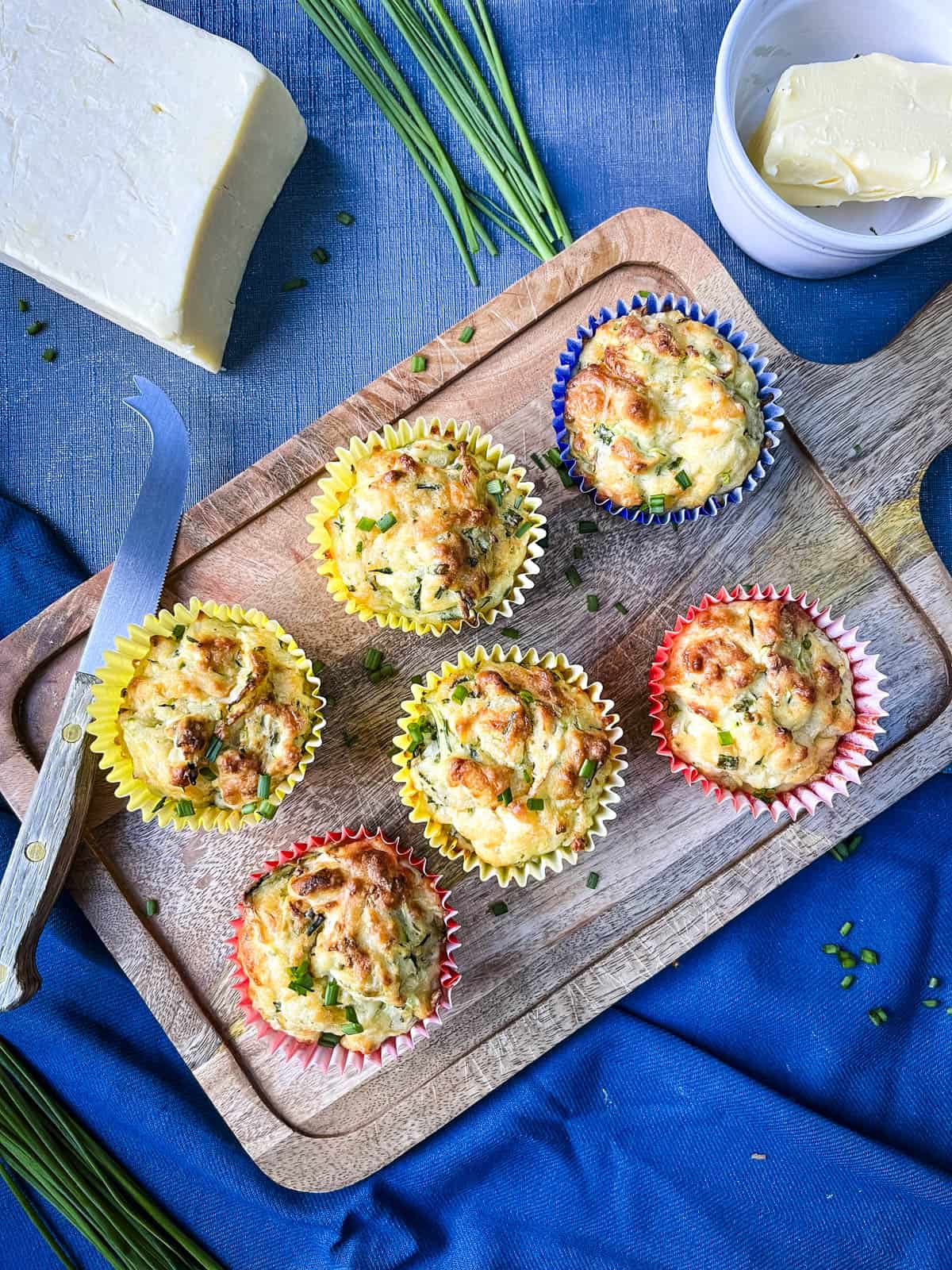cheese and courgette muffins with chives on wooden board with butter and cheese to side.