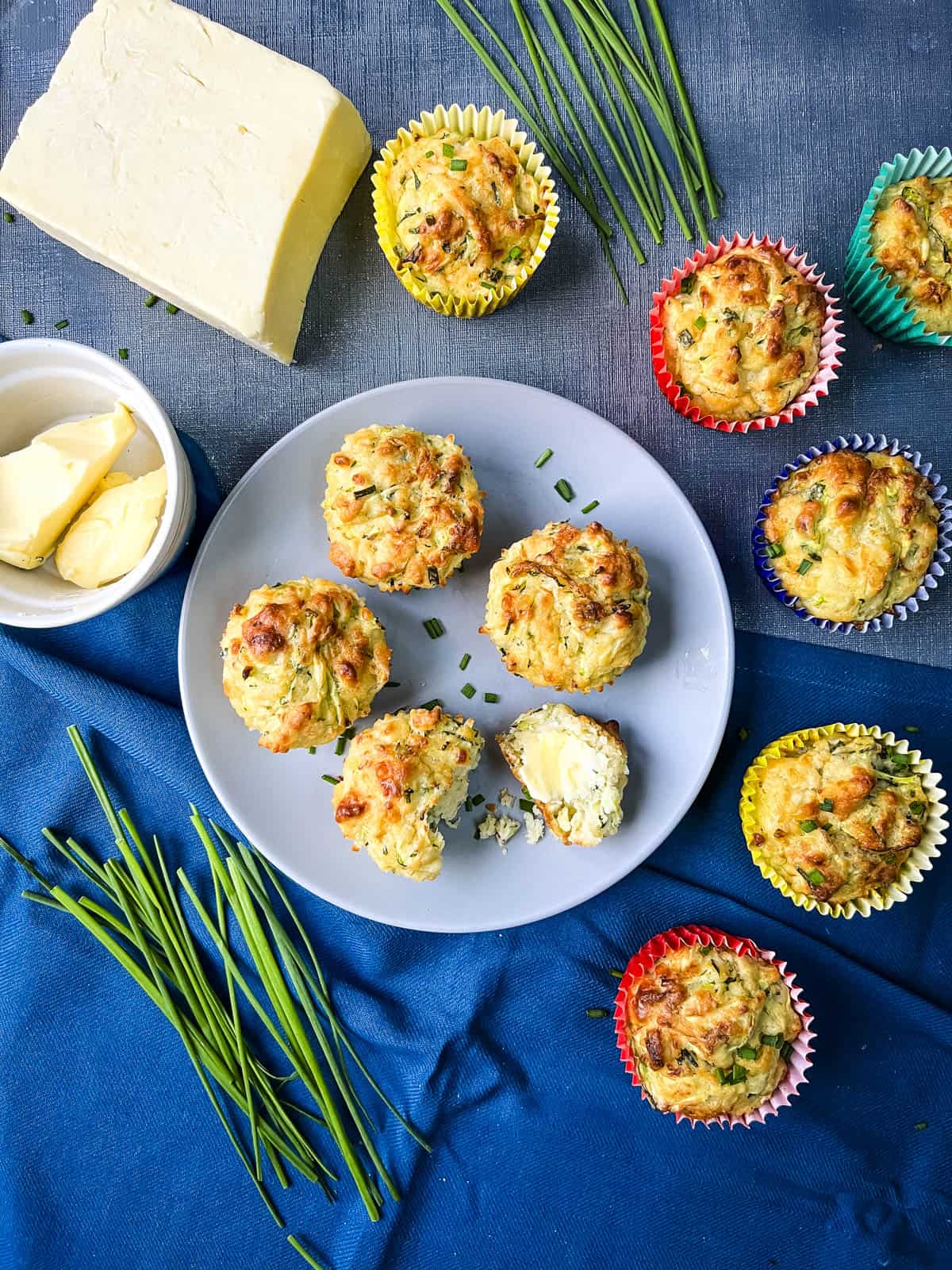cheese and courgette muffins on a plate with more to the side and fresh chives, cheddar cheese and butter in a bowl.