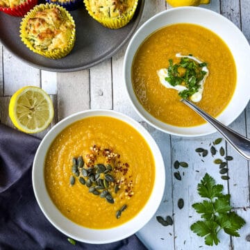 two bowls of carrot and cumin soup topped with pumpkin seeds and fresh parsley with carrot and courgette muffins to side