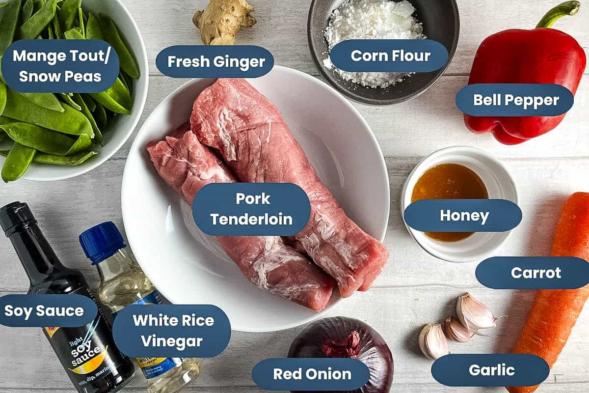 labelled ingredients for sticky pork stir fry, pork tenderloin, mangetout, red pepper, fresh ginger, cornflour, honey, carrot, garlic, red onion, white rice vinegar, soy sauce.