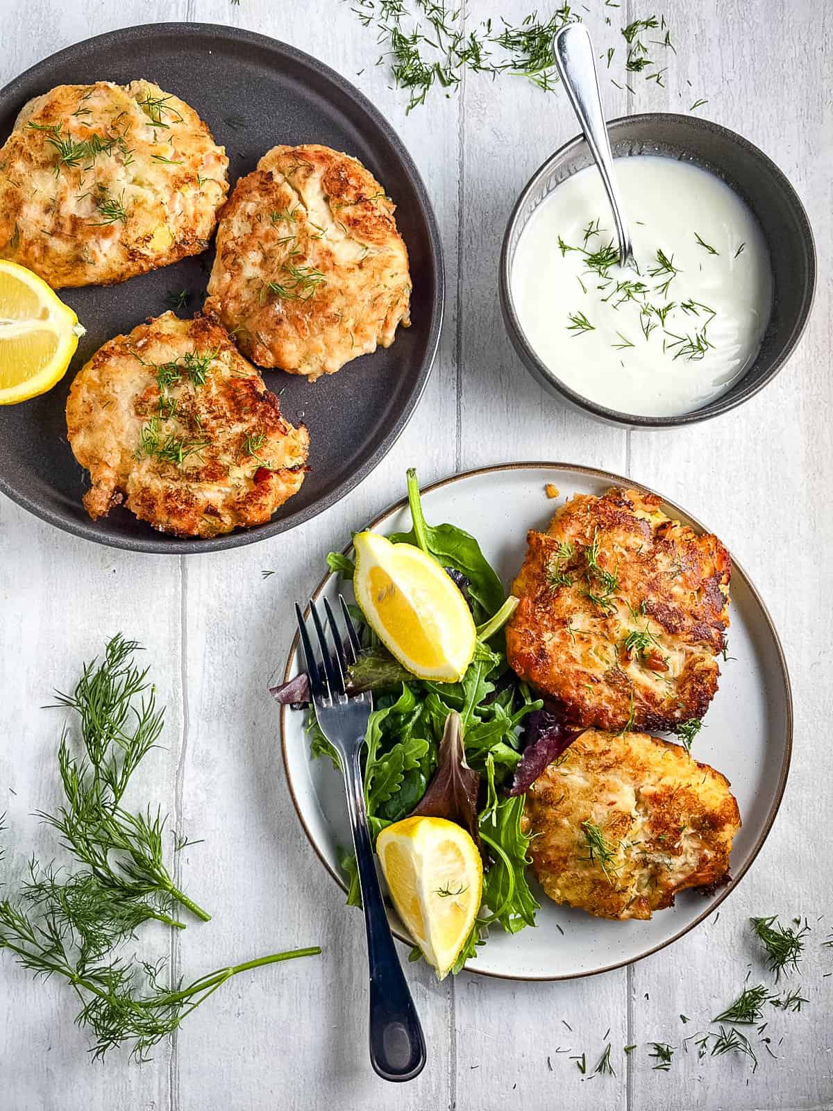 cokked salmon fishcakes on a plate with green salad and lemon wedges with a side of white sauce and plate of more fishcakes.