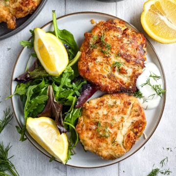 two salmon fishcakes on a plate with green salad and lemon wedges.