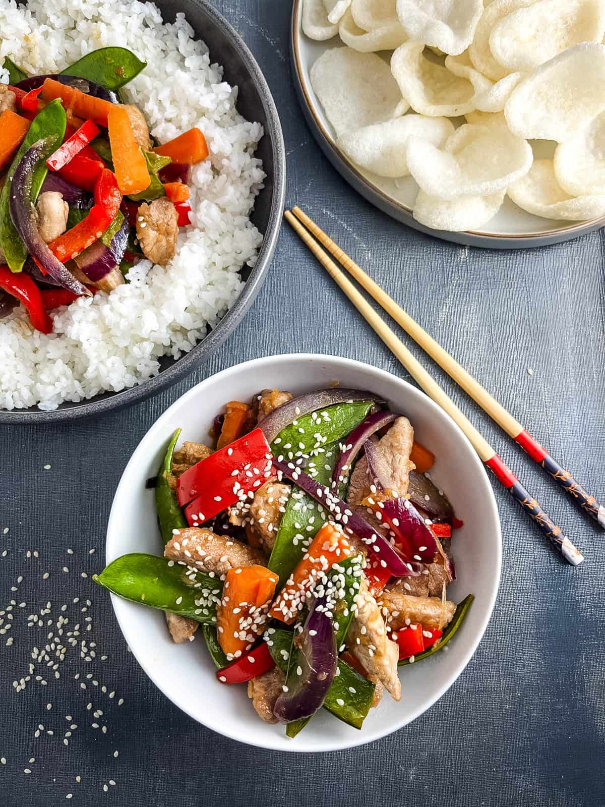 sticky pork stir fry in a bowl topped with sesame seeds with a side of prawn crackers.