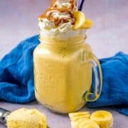 glass mug of banana milkshake topped with whipped cream, banana slices and caramel sauce.