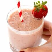 hand holding glass of strawberry banana smoothie with red and white paper straw.