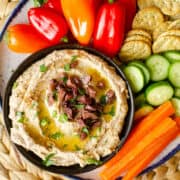 bowl of olive hummus served with fresh carrot sticks, cucumber, peppers and crackers.